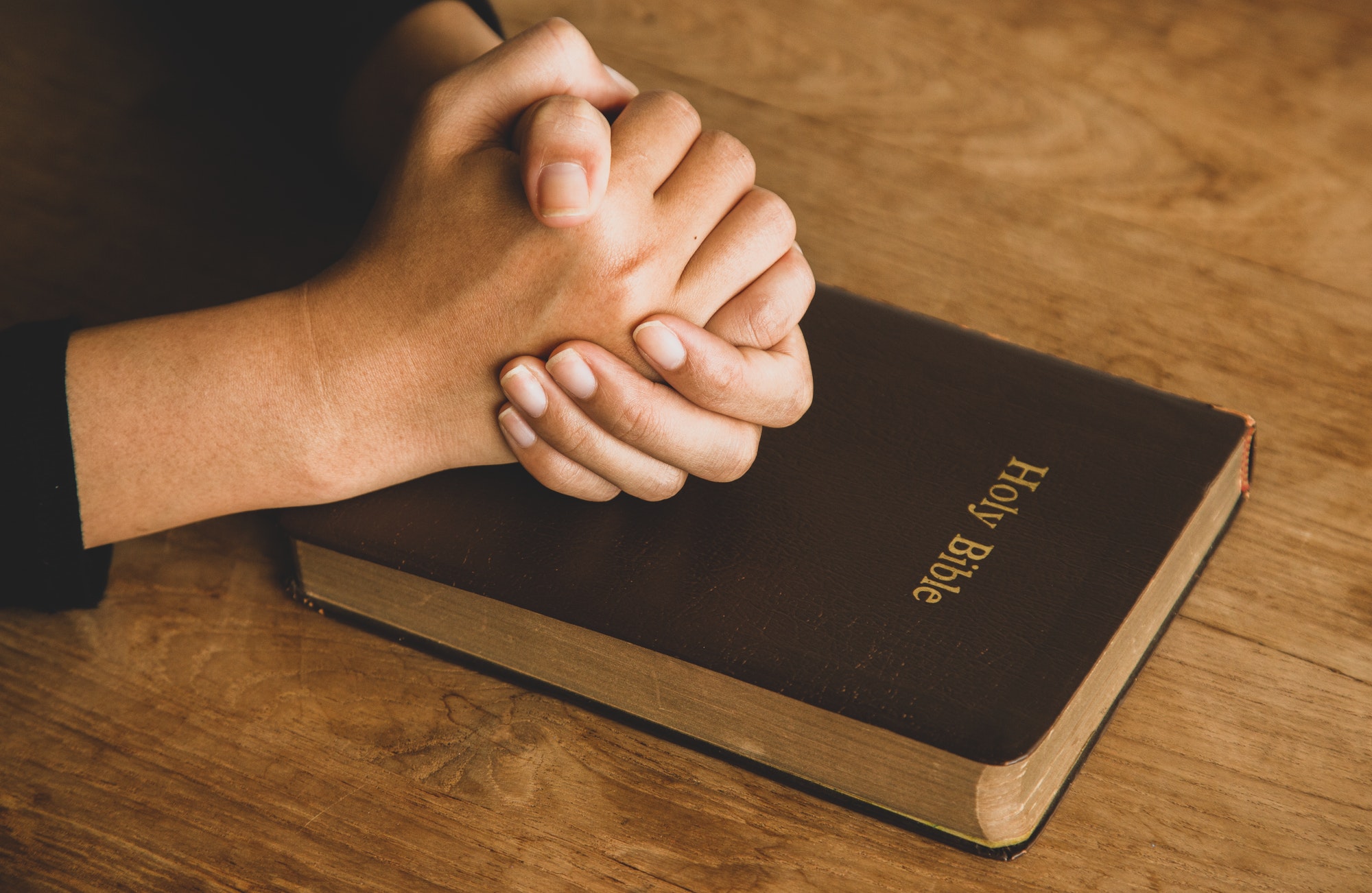 Woman with Bible praying, hands clasped together on her Bible on wooden table